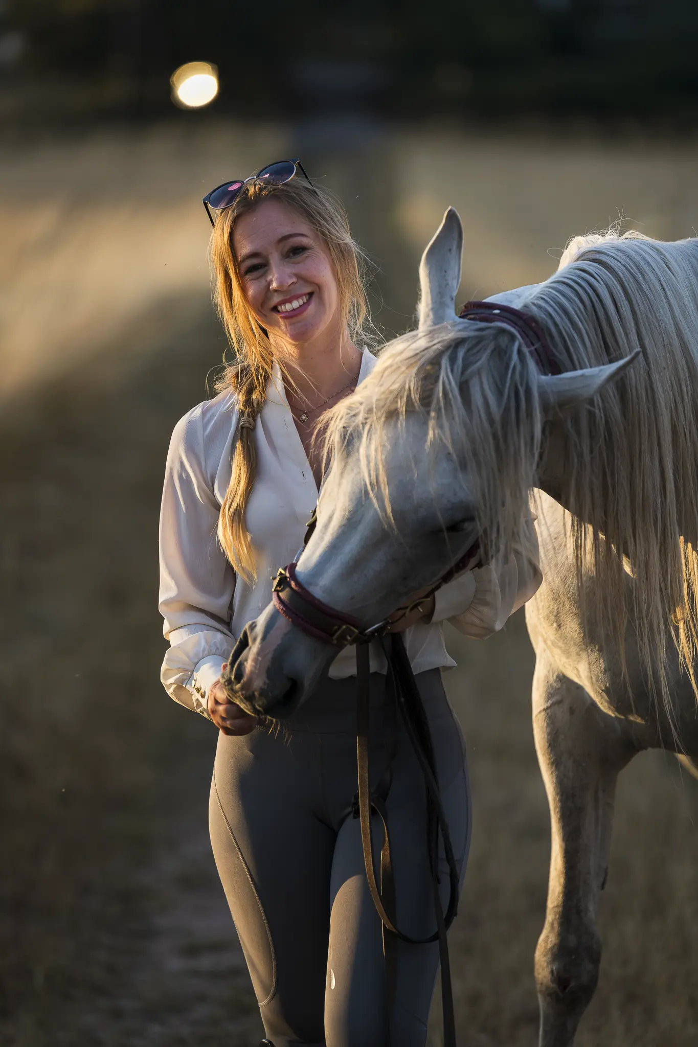Ein vertikales Outdoor-Porträt einer lächelnden Frau mit blondem Zopf, die in der Abendsonne (Golden Hour) neben einem weißen Pferd steht. Sie trägt eine elegante weiße Bluse, eine Sonnenbrille auf dem Kopf und graue Reithosen. Das Pferd senkt den Kopf zärtlich zu ihr, während das warme Gegenlicht die Konturen beider Motive golden hervorhebt. Der Hintergrund ist in einer stimmungsvollen Unschärfe gehalten, die den Fokus ganz auf die harmonische Verbindung zwischen Mensch und Tier lenkt.