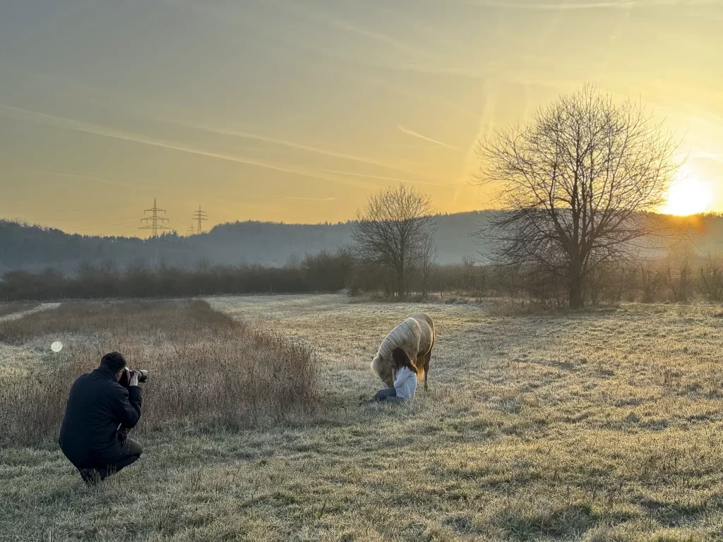 Behind-the-Scenes-Aufnahme des Tierfotografen bei der Arbeit auf einer winterlichen Wiese während des Sonnenuntergangs. Der Fotograf kniet im Vordergrund mit einer professionellen Kamera und Tele-Objektiv. Im Hintergrund sitzt eine junge Frau im sanften Gegenlicht neben einem hellen Pferd auf dem Boden. Die Szene ist in warmes, goldenes Licht getaucht, im Hintergrund sind Hügel und Bäume in einer leichten Dunstschicht zu sehen. Fokus auf authentische Pferdefotografie und professionelles Equipment in der Golden Hour.