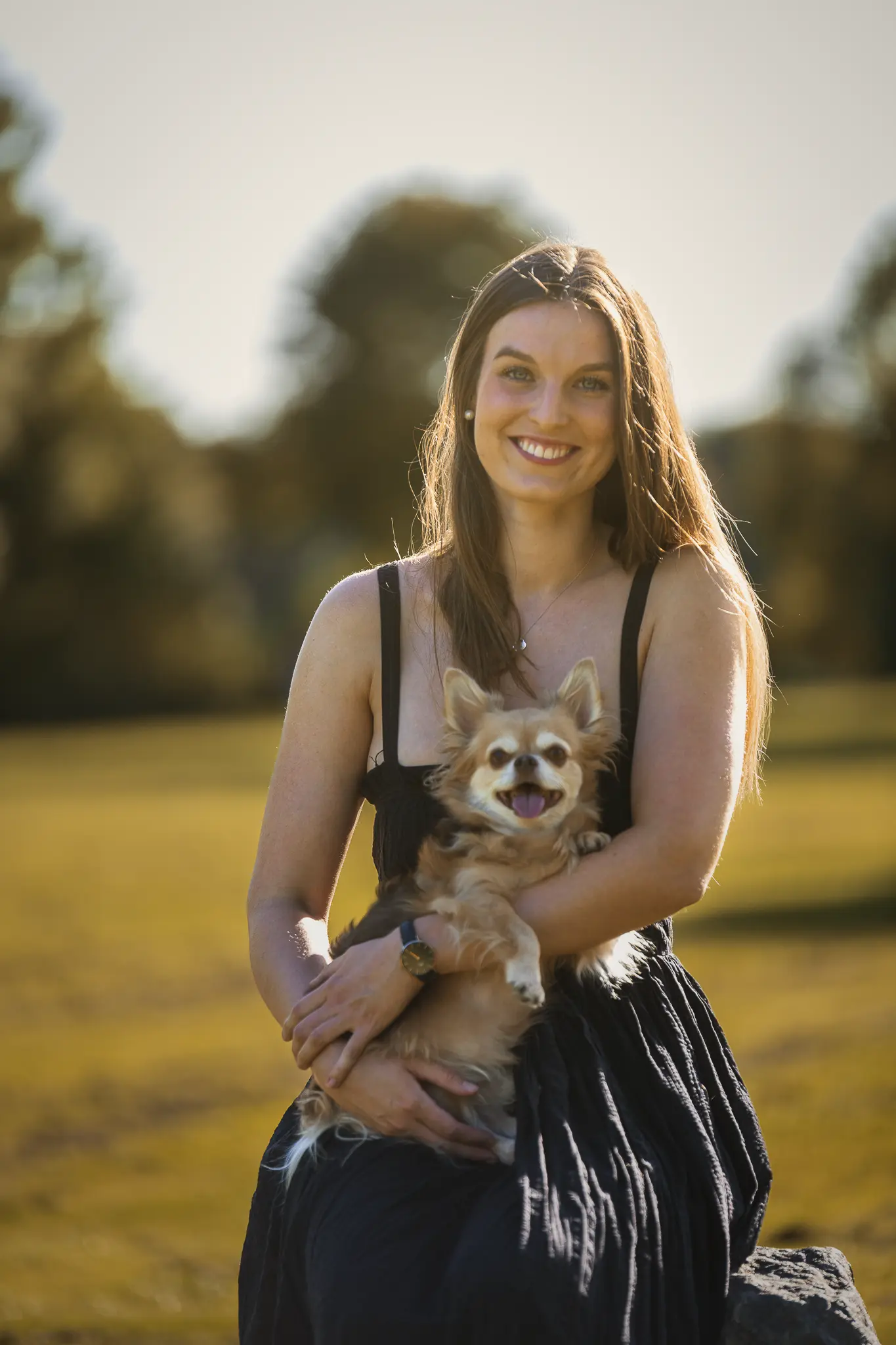 Ein herzliches Outdoor-Porträt einer lächelnden Frau, die einen kleinen, langhaarigen Chihuahua liebevoll im Arm hält. Das Foto wurde im warmen Gegenlicht auf einer herbstlichen Wiese aufgenommen, wodurch eine sanfte goldene Aura um die Motive entsteht. Der Hintergrund ist in einer weichen Unschärfe gehalten. Professionelle Hundefotografie und Tierporträts im Raum Frankfurt und Hattersheim von René Hering.
