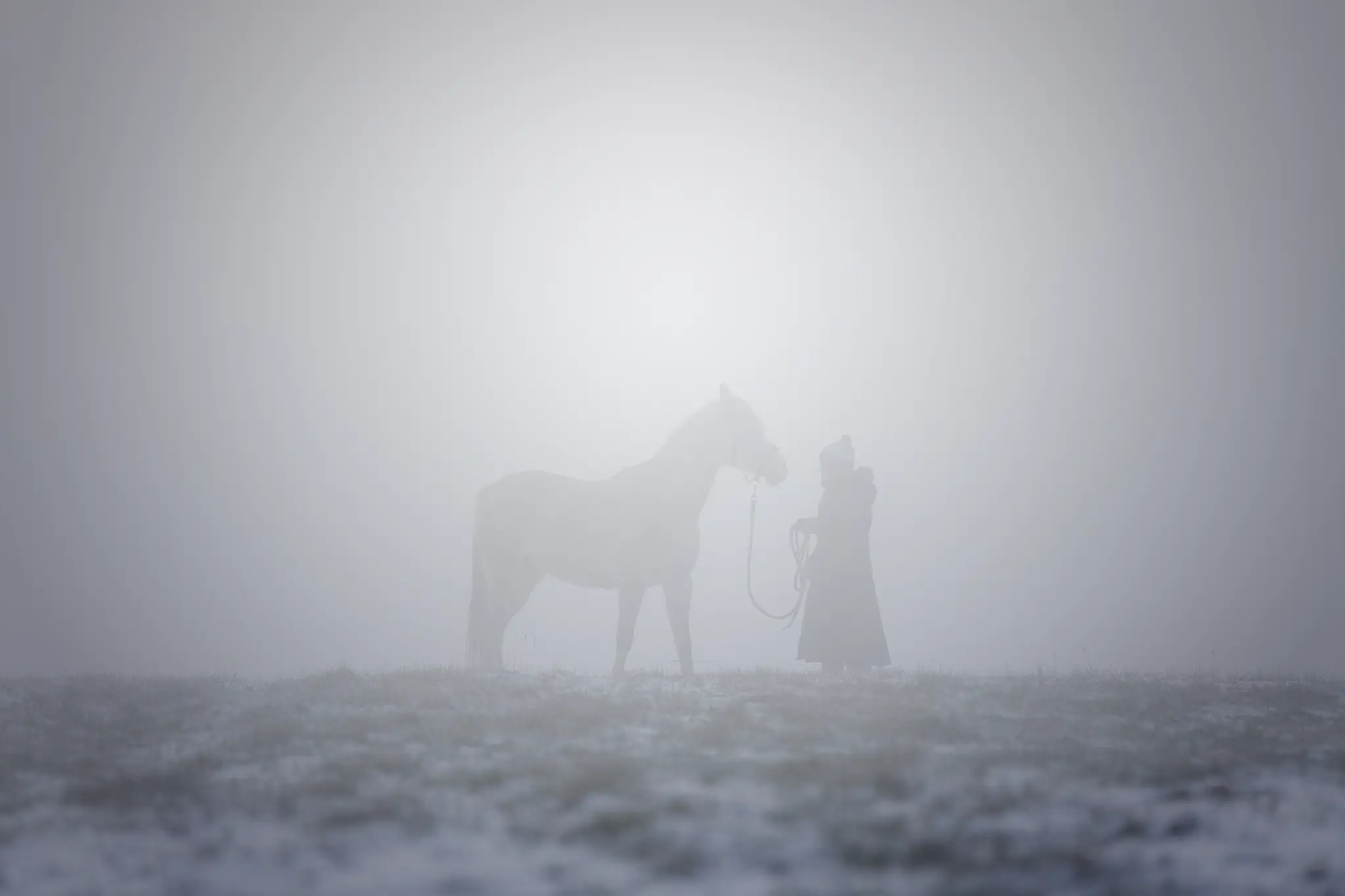 Eine minimalistische Fine-Art-Aufnahme bei dichtem Nebel. Man sieht die dunklen Silhouetten einer Person und eines Pferdes auf einer leicht verschneiten oder frostigen Wiese. Das Licht bricht sanft durch den weißen Nebel und lässt die Konturen verschwimmen. Eine sehr ruhige, atmosphärische Darstellung der Verbundenheit zwischen Mensch und Tier im Winter. Künstlerische Pferdefotografie von René Hering.