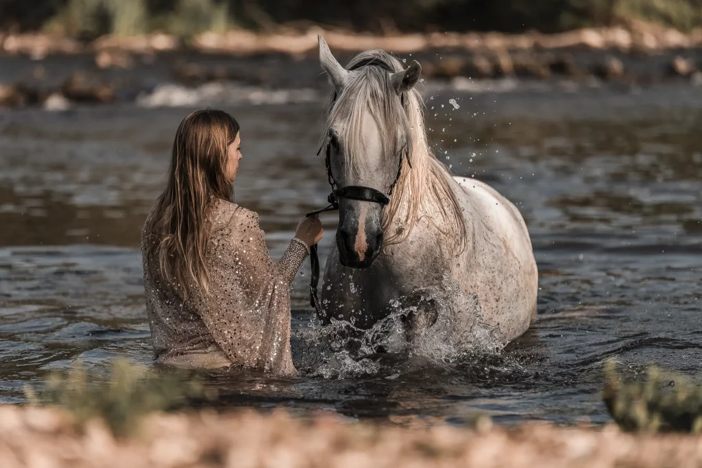 Ein künstlerisches Fine-Art-Porträt einer Frau in einem glitzernden, hellen Kleid und eines weißen Pferdes, die gemeinsam in einem Fluss stehen. Das Pferd wirbelt mit den Vorderhufen Wasser auf, wodurch dynamische Wasserspritzer entstehen. Die Szene ist in gedeckten, erdigen Tönen bearbeitet, was eine elegante und zeitlose Atmosphäre schafft. Professionelle Tierfotografie im Wasser, aufgenommen von René Hering im Rhein-Main-Gebiet.