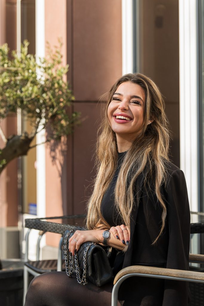 Authentisches Business-Porträt einer Frau im schwarzen Blazer, die herzlich lachend an einem Tisch im Freien sitzt. Im Hintergrund sind ein Olivenbaum und eine moderne Gebäudefassade in der Sonne zu sehen.