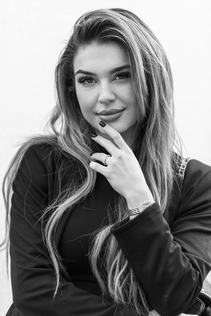 Klassisches Schwarz-Weiß-Businessporträt einer jungen Frau mit langen Haaren, die ihre Hand leicht ans Kinn legt und direkt in die Kamera blickt. Sie trägt einen dunklen Blazer und eine elegante Armbanduhr.