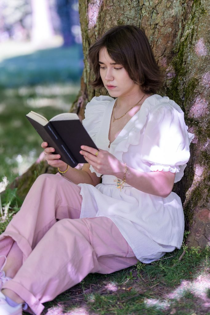 Junge Frau in weißer Bluse sitzt im weichen Sommerlicht an einem Baumstamm und liest in einem Buch.
