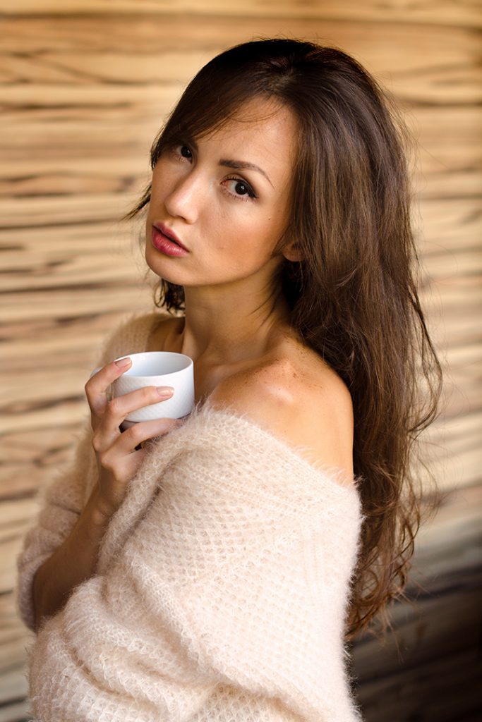 Eine junge Frau mit langen braunen Haaren und einem hellen, flauschigen Strickpullover hält eine weiße Kaffeetasse und blickt direkt in die Kamera.