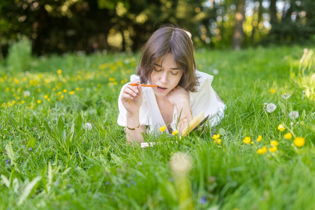 Eine junge Frau liegt im hohen Gras auf einer Sommerwiese und überlegt mit einem Stift im Mund über ihrem Notizbuch.