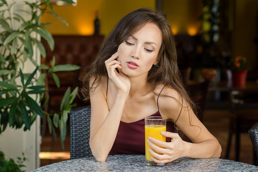 Eine junge Frau sitzt nachdenklich an einem Marmortisch in einem Café und hält ein Glas Orangensaft in der Hand.